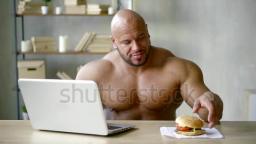 Huge weightlifter biting big BURGER sitting behind laptop indoor